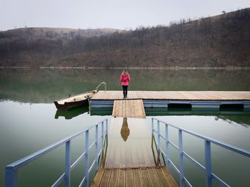 Woman with pink coat and backpack walking down a wooden pontoon towards a lake in autumn