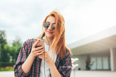 Beautiful young woman listening music on mobile phone against sky