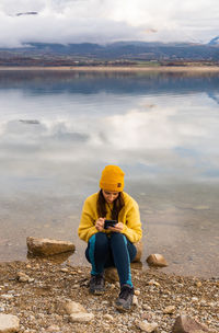 Woman using phone sitting on the lake shore with the reflection of the mountain over the lake