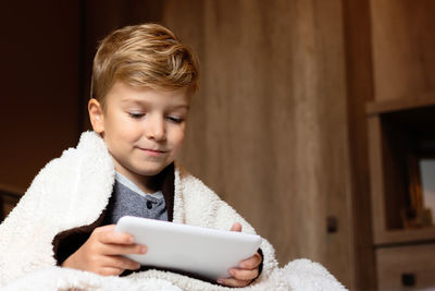 Small boy wrapped in a blanket using digital tablet at home.