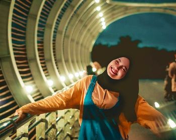 Portrait of smiling young woman with arms outstretched standing against built structure