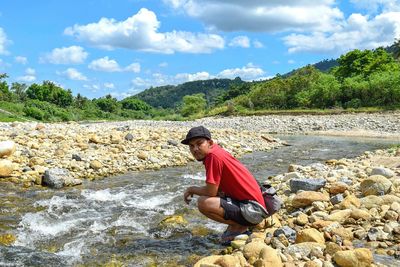 Side view of young man crouching at riverbank against sky