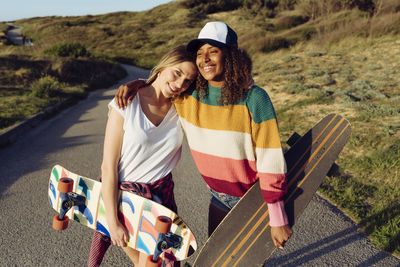 Two beautiful multiethnic cheerful women skaters holding skate boards while walking in the middle of a road on a sunny day