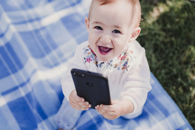 High angle portrait of smiling baby girl holding phone on picnic blanket