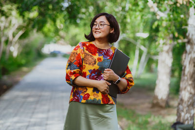 A smiling asian woman with glasses holding a laptop in a sunny park.