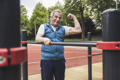 Happy senior man flexing muscles standing by gymnastics bar