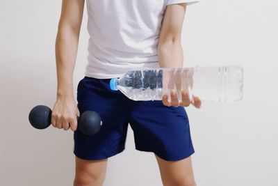 Midsection of man holding dumbbell and water bottle against white background