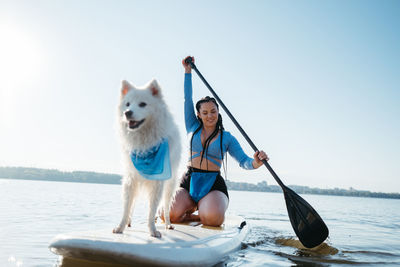 Woman paddleboarding with her pet on city lake, snow-white japanese spitz dog standing on sup board