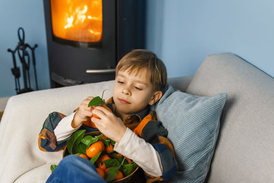 Boy peels and eats a tangerine, sitting in an armchair by the fireplace.