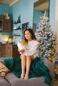 Young woman in cozy home clothes is resting on the sofa in the living room, decorated for christmas