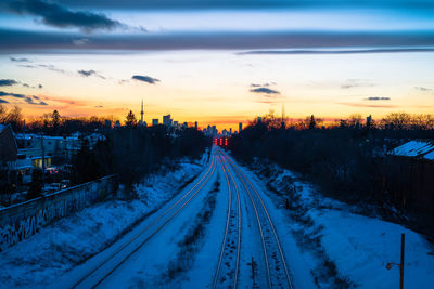 Snow covered railroad tracks in city against sky during sunset