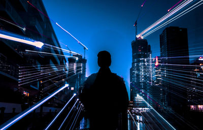 Rear view of man standing by illuminated buildings against sky at night