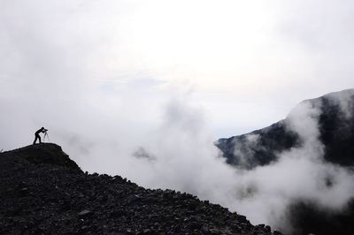 Silhouette man photographing on mountain against sky during foggy weather