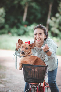 Cheerful girl with dog in basket of bicycle standing outdoors