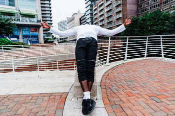 REAR VIEW OF MAN STANDING ON FOOTPATH BY STREET AGAINST BUILDINGS