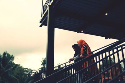 Low angle view of man standing by railing against orange sky