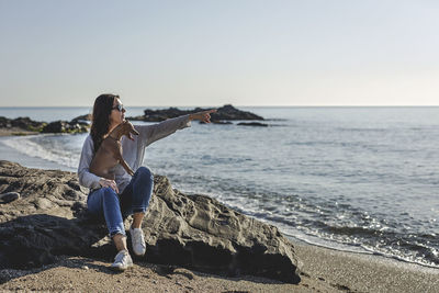 Full length of woman and dog looking at sea while sitting on rock at beach