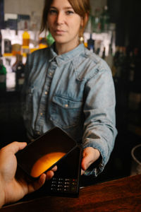 Crop anonymous client with making contactless payment by modern smartphone on terminal in bar near female bartender looking at camera