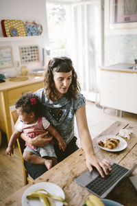 Mother using laptop while sitting with daughter at table in house