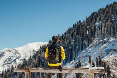 Rear view of man sitting on park bench against mountains and sky during winter