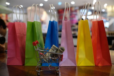 Close-up of colorful shopping bags with paper currencies in miniature shopping cart on wooden table