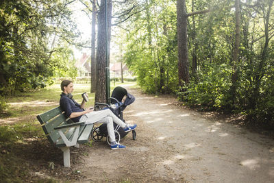 Full length of man using smart phone while sitting by baby sleeping in carriage at park