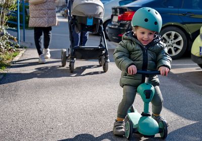 Little toddler on scooter and his parents with baby stroller walking down the street