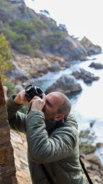 Side view of man photographing while sitting on rock