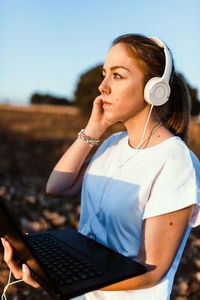 Side view of woman listening while using laptop on land against sky