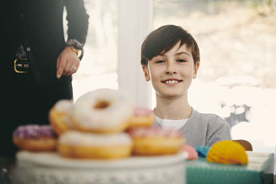 Portrait of smiling boy sitting by grandmother at dining table with donuts in foreground