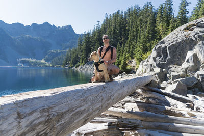 Man with dog crouching on fallen tree by alpine lake