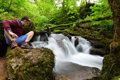 Young man sitting on rock by waterfall in forest