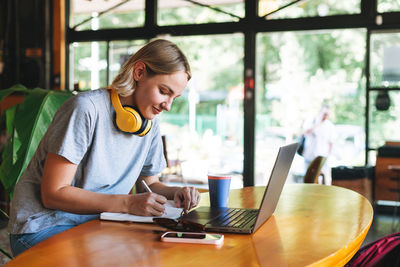 Young smiling blonde woman freelancer with yellow headphones working on notebook on table at cafe