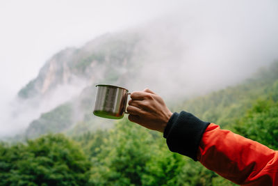 Midsection of person holding drinking cup against foggy mountains