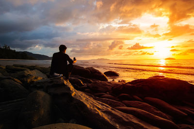 Rear view of man sitting on rock at beach against sky during sunset