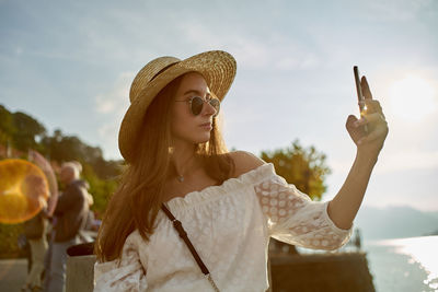 Portrait of young woman holding hat against sky