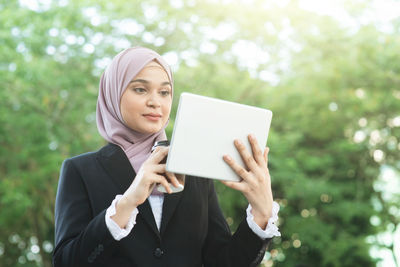 Young woman using digital tablet while holding coffee cup against trees