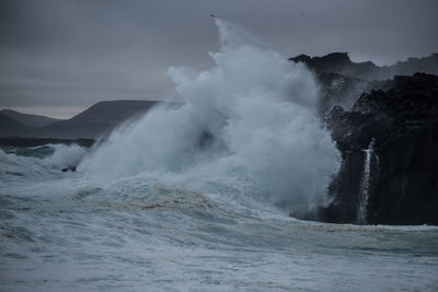 Waves splashing on rocks at shore against sky