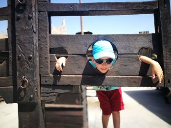 Boy in blue cap and sunglasses peeking head from circle of wooden structure
