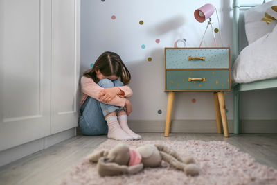 Sad little girl sitting on the floor of her bedroom with stuffed toy lying