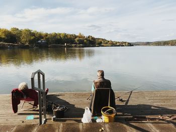 Rear view of man sitting on table by lake against sky