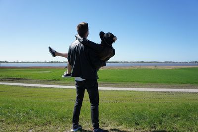 Rear view of boyfriend carrying girlfriend while standing on grassy field against sky during sunny day