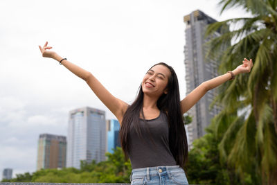 Young woman with arms raised standing in city against sky