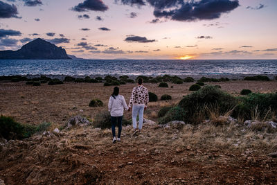 Rear view of people standing on beach against sky during sunset