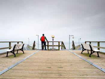 Tourist with red backpack on wooden pier above sea. man in trekking suit in harbor in dark rainy day