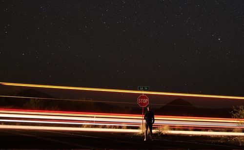 Man standing by light trails on road against sky at night