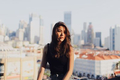 Young woman with long hair looking away while sitting against modern buildings in city