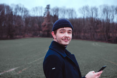 Portrait of man wearing knit hat using phone while standing on field
