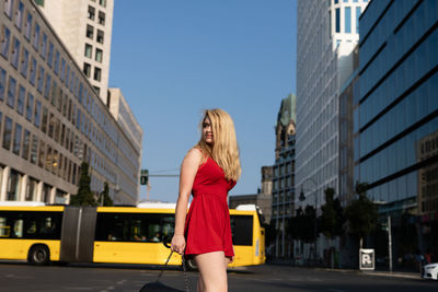 Woman looking away while standing on road against bus and buildings in city
