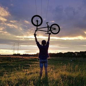 Rear view of man lifting bicycle on field against sky